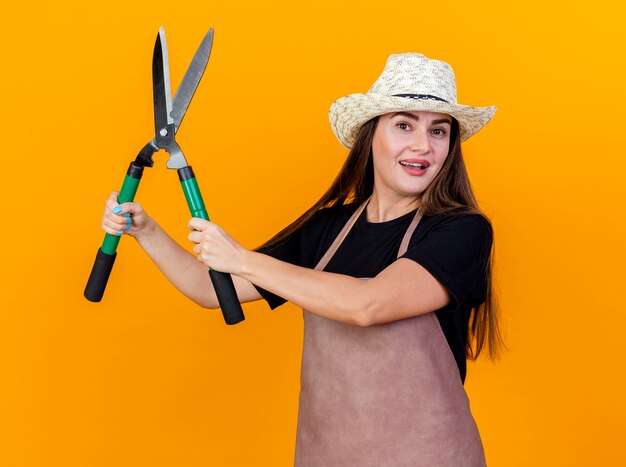 Sorridente bella ragazza giardiniere che indossa uniforme e cappello da giardinaggio sollevando clippers isolato su sfondo arancione
