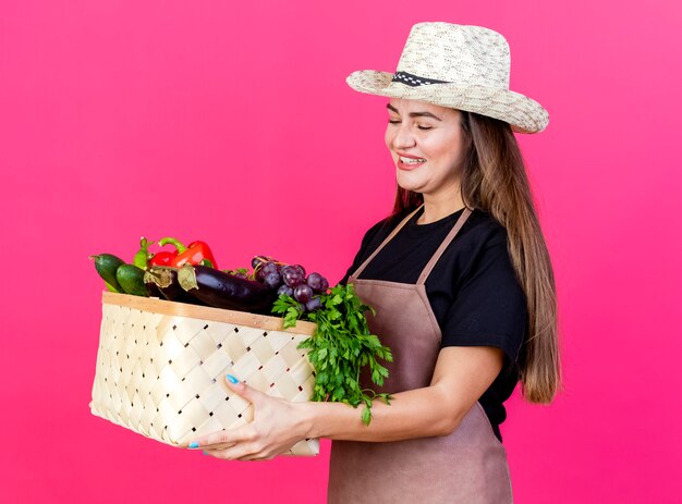 Sorridente bella ragazza giardiniere in uniforme che indossa cappello da giardinaggio tenendo e guardando il cesto di verdure isolato su sfondo rosa