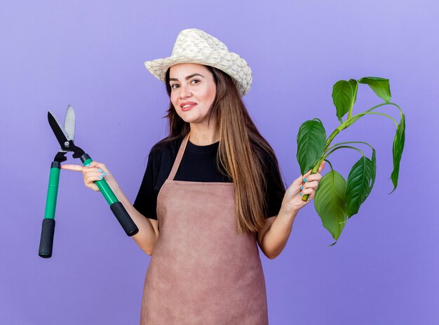 Sorridente bella ragazza giardiniere in uniforme che indossa cappello da giardinaggio tenendo la pianta con le forbici e diffondendo le mani isolate sull'azzurro