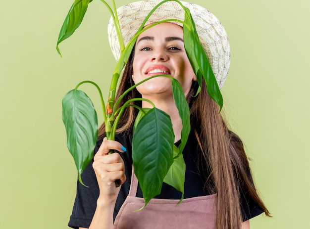 Sorridente bella ragazza giardiniere in uniforme che indossa cappello da giardinaggio viso coperto con pianta isolata su verde oliva