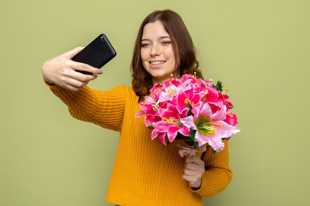 Sorridente bella ragazza il giorno delle donne felici tenendo il bouquet prendere un selfie isolato su parete verde oliva