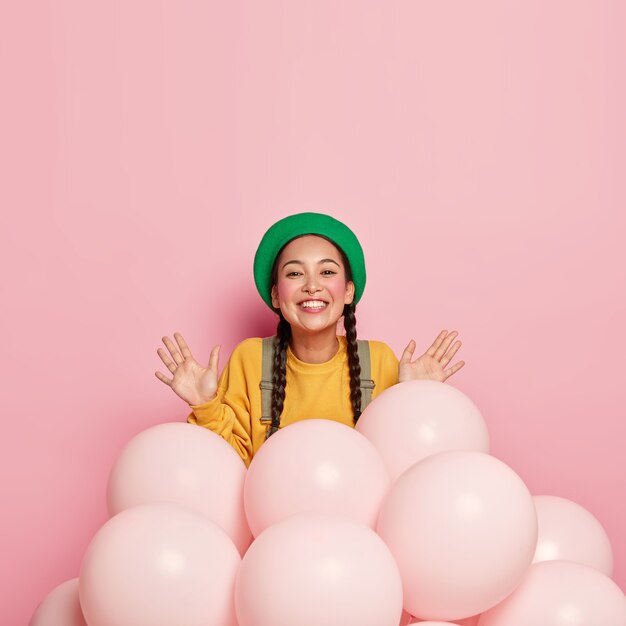 Sorridente bella signora asiatica tiene le palme sollevate vicino a palloncini di elio, essendo di buon umore, indossa berretto verde e maglione casual giallo, decora la stanza per eventi speciali