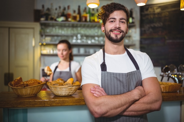 Sorridente cameriere in piedi con le braccia incrociate in caffetteria ©