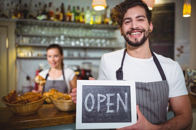 Sorridente cameriere mostrando ardesia con segno aperto in caffetteria ©