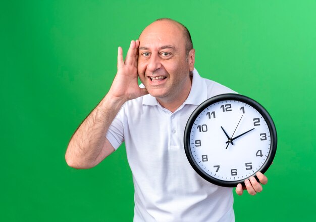 Sorridente casual uomo maturo tenendo l'orologio da parete e mettendo la mano sulla guancia isolata sulla parete verde