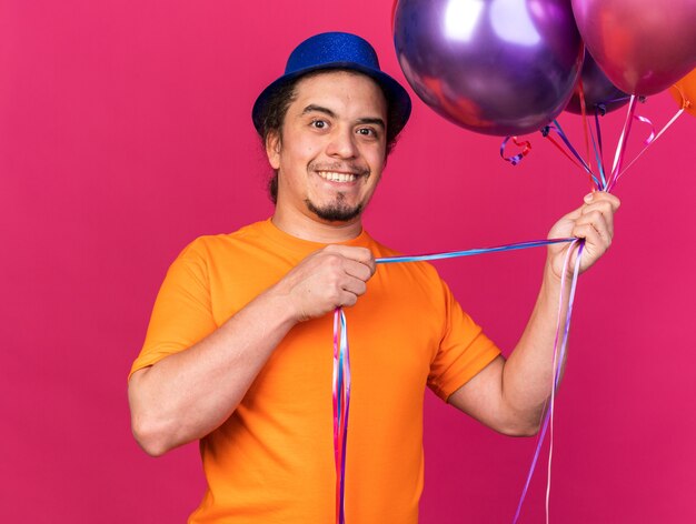 Sorridente cercando fotocamera giovane uomo che indossa cappello da festa tenendo palloncini isolati su parete rosa