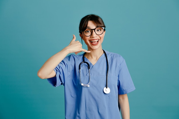 Sorridente che mostra gesto di chiamata telefonica giovane dottoressa che indossa uno stetoscopio fitth uniforme isolato su sfondo blu