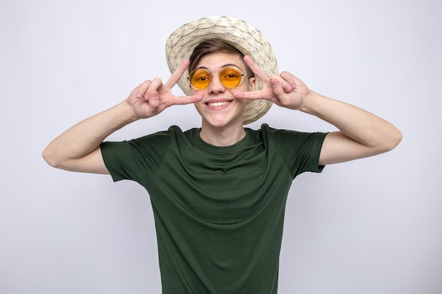 Sorridente che mostra gesto di pace giovane bel ragazzo che indossa un cappello con gli occhiali isolati sul muro bianco