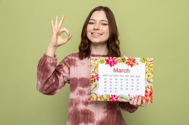 Sorridente che mostra gesto ok bella ragazza il giorno della donna felice che tiene il calendario isolato sul muro verde oliva