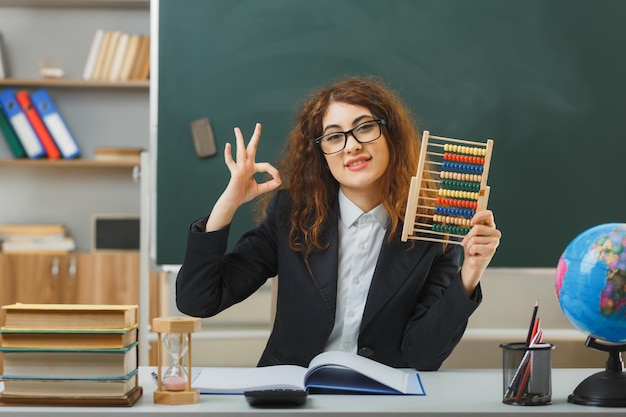 sorridente che mostra gesto ok giovane insegnante femminile che indossa occhiali con in mano abaco seduto alla scrivania con strumenti scolastici in classe