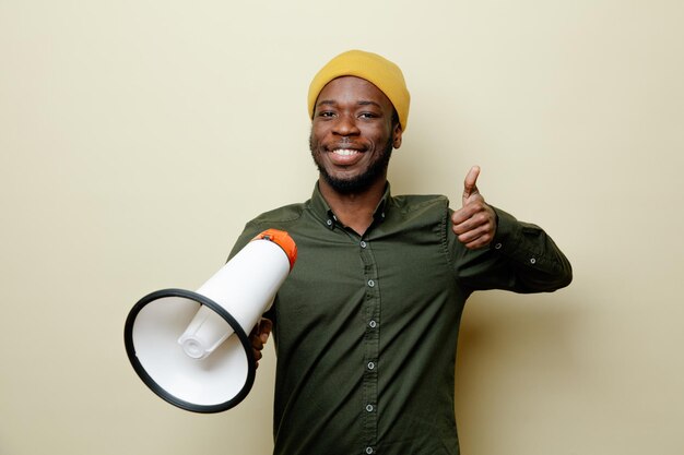 Sorridente che mostra i pollici in su giovane maschio afroamericano con il cappello che indossa una camicia verde che tiene l'altoparlante isolato su sfondo bianco