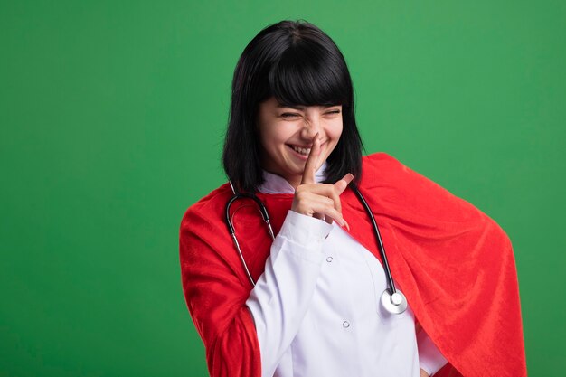 Sorridente con gli occhi chiusi giovane ragazza del supereroe che indossa uno stetoscopio con veste medica e mantello che mostra gesto di silenzio isolato sulla parete verde