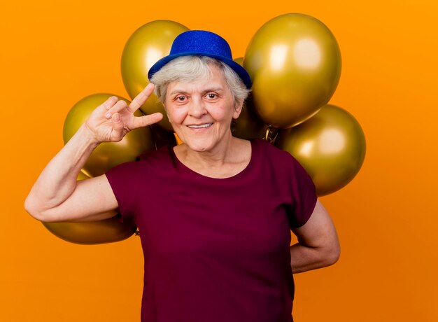 Sorridente donna anziana che indossa il cappello del partito gesti il segno della mano di vittoria e tiene palloncini di elio