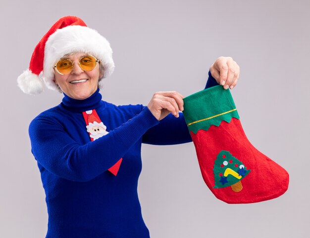 Sorridente donna anziana in occhiali da sole con cappello da Babbo Natale e cravatta di Babbo Natale che tiene calza di Natale isolata sul muro bianco con spazio di copia