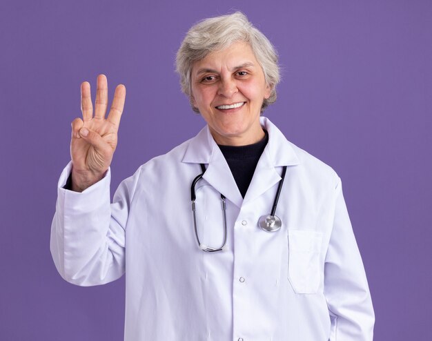 Sorridente donna anziana in uniforme da medico con stetoscopio che gesturing tre con le dita