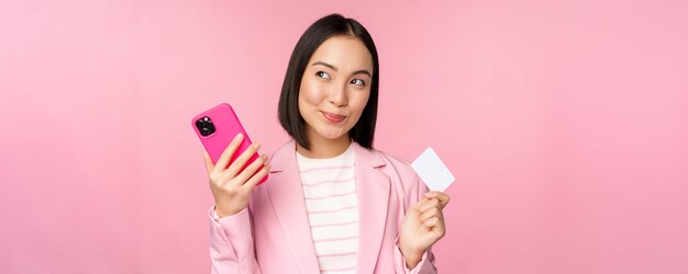 Sorridente donna asiatica aziendale signora in tuta pensando tenendo smartphone e carta di credito piano per acquistare smth shopping online con telefono cellulare sfondo rosa