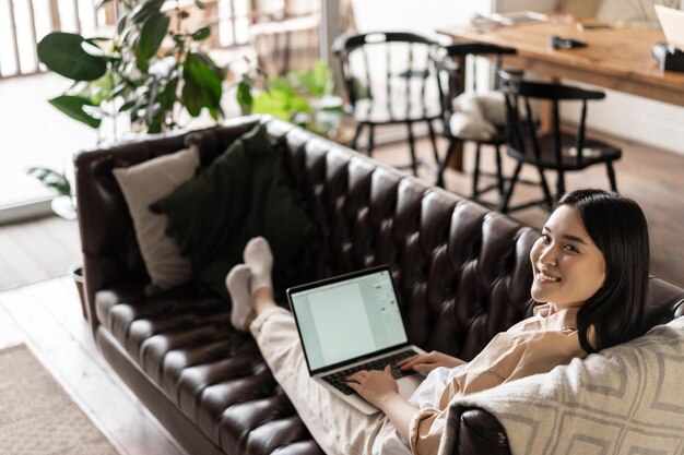 Sorridente donna asiatica che lavora freelance da casa che riposa sul divano con un computer portatile che sembra felice ...