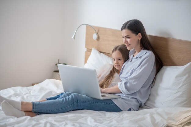 Sorridente donna attraente e il suo bambino carino e tranquillo seduto sul letto davanti al computer notebook