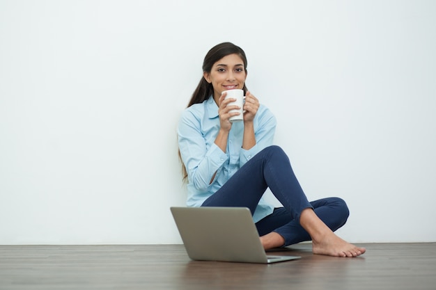 Sorridente donna bevendo tè sul pavimento con il computer portatile