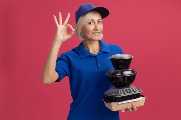 Sorridente donna bionda di mezza età di consegna in uniforme blu e berretto che tiene in mano un pacchetto di alimenti di carta e contenitori per alimenti che fanno segno ok isolato sul muro rosa
