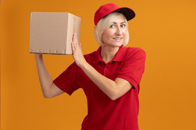 Sorridente donna bionda di mezza età in uniforme rossa e berretto che tiene una scatola di cartone vicino alla testa