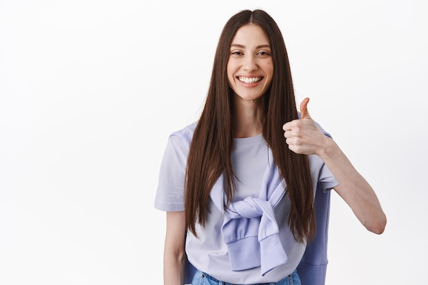 Sorridente donna bruna che mostra pollice in su, annuisce in segno di approvazione, supporta una buona scelta, loda un ottimo lavoro, cosa eccellente, in piedi su un muro bianco