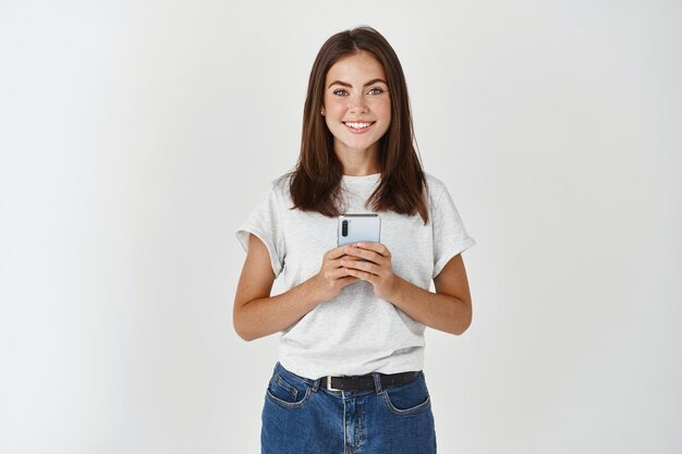 Sorridente donna bruna che tiene smartphone e guarda davanti, in piedi in una maglietta casual sul muro bianco.