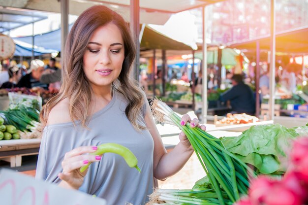 Sorridente donna con verdure al negozio di mercato. Donna che sceglie verdure fresche nel mercato verde. Ritratto di bella giovane donna che sceglie verdure a foglia verde