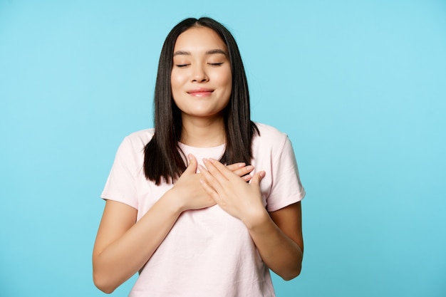 Sorridente donna coreana che sogna, tenendosi per mano sul cuore e chiudendo gli occhi, custodisce il ricordo nella sua anima, in piedi su sfondo blu.