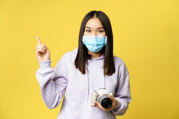 Sorridente donna coreana in maschera medica, tenendo la macchina fotografica, chiedendo di scattare una foto, indicando qualcosa su uno spazio vuoto, sfondo giallo