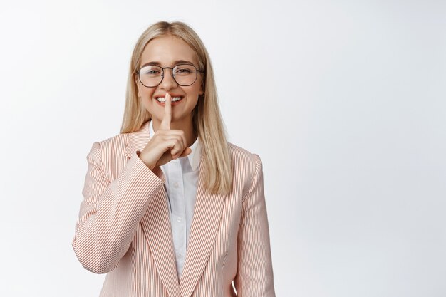 Sorridente donna d'affari che mostra il segno del silenzio, mantenendo un segreto, fa un gesto shh, in piedi in tuta e occhiali su bianco