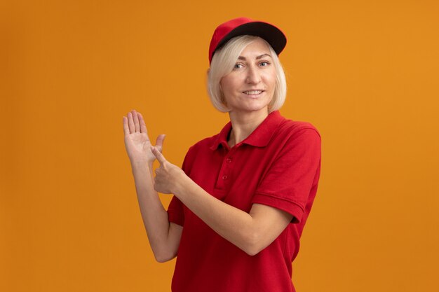 Sorridente donna di mezza età bionda consegna in uniforme rossa e berretto in piedi in vista di profilo che mostra la mano vuota e indicando a portata di mano isolata sulla parete arancione con spazio di copia