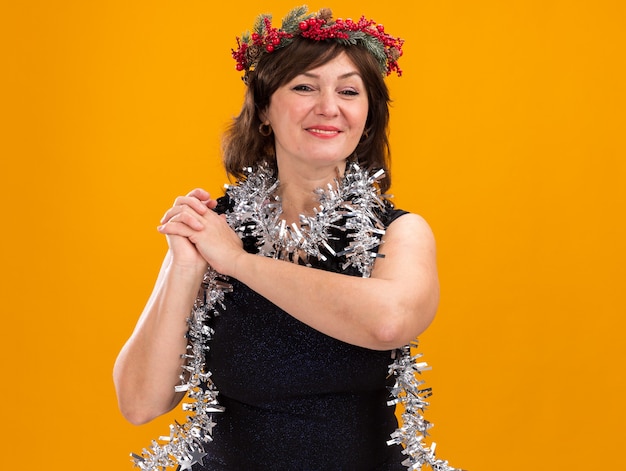 Sorridente donna di mezza età che indossa la testa di Natale ghirlanda e tinsel ghirlanda intorno al collo tenendo le mani insieme guardando la telecamera isolata su sfondo arancione con spazio di copia