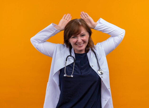 Sorridente donna di mezza età medico indossa abito medico e stetoscopio mettendo le mani sopra la testa sulla parete arancione isolata