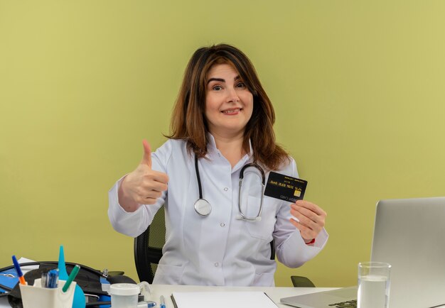 Sorridente donna di mezza età medico indossa abito medico e stetoscopio seduto alla scrivania con strumenti medici e laptop tenendo la carta di credito che mostra il pollice in alto isolato