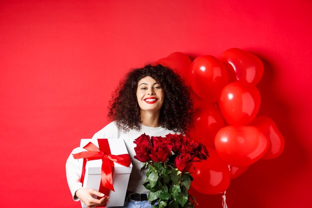 Sorridente donna felice che tiene scatola con regalo e rose rosse dal fidanzato che celebra San Valentino st...