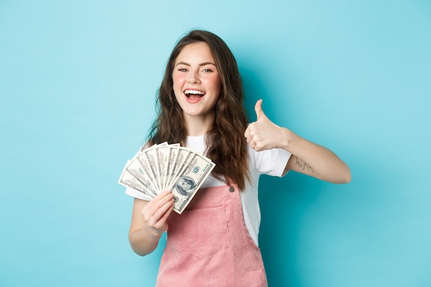 Sorridente donna felice che tiene soldi, banconote da un dollaro e mostra il pollice in su, raccomanda un prestito in contanti veloce e sembra soddisfatta, in piedi su sfondo blu