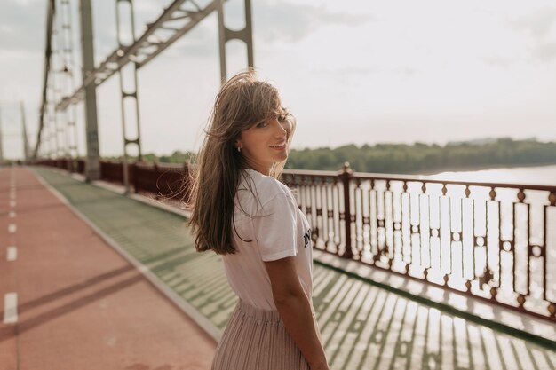 Sorridente donna felice con lunghi capelli ondulati che indossa maglietta bianca e gonna sta camminando all'aperto sul ponte con vista sul mare alla luce del sole del mattino