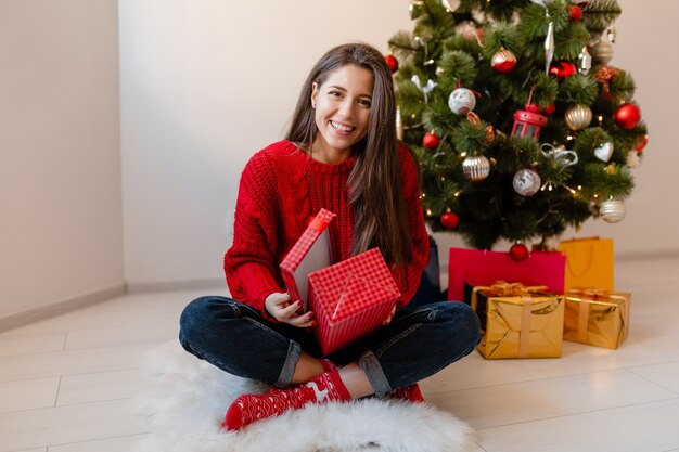 Sorridente donna graziosa eccitata in maglione rosso seduto a casa all'albero di Natale disimballaggio regali e scatole regalo