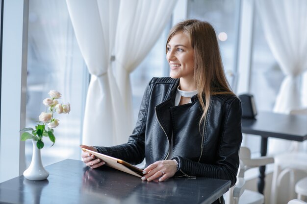 Sorridente donna seduta in un ristorante con una tavoletta guardando fuori dalla finestra