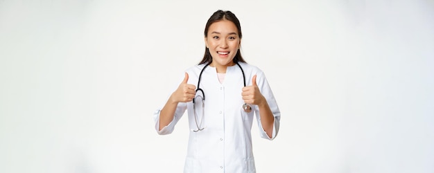 Sorridente dottoressa asiatica che indossa un abito medico che mostra i pollici in su lieto di consigliare prodotti per la salute