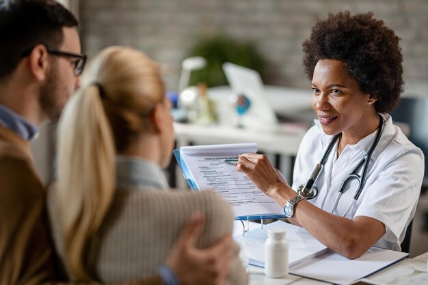 Sorridente dottoressa nera che parla con una coppia mentre analizza i loro referti medici durante le consultazioni in clinica