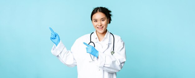 Sorridente felice dottoressa o infermiera che punta le dita a sinistra indossando uniforme medica e guanti showin