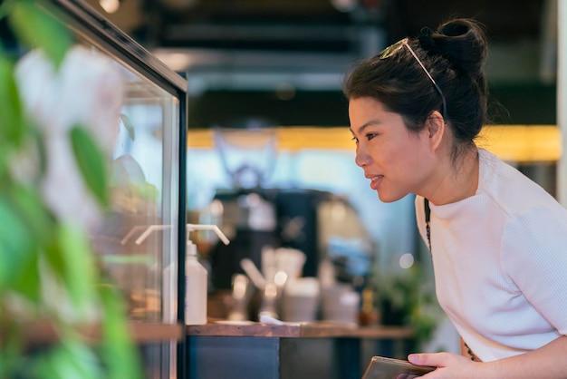 Sorridente femmina asiatica donna adulta felicità allegra selezionando cioccolatini pregiati torta e confetteria al caffè armadio display sfondo del caffè