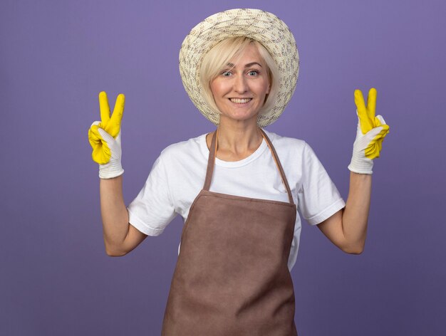 Sorridente giardiniere bionda di mezza età donna in uniforme che indossa cappello e guanti da giardinaggio facendo segno di pace