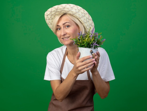 Sorridente giardiniere bionda di mezza età donna in uniforme che indossa un cappello che tiene vaso di fiori