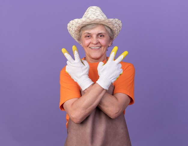 Sorridente giardiniere donna anziana che indossa cappello e guanti da giardinaggio che si incrociano le mani gesticolando segno di vittoria victory