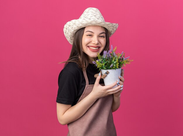 Sorridente giardiniere femmina caucasica che indossa cappello da giardinaggio con fiori in vaso di fiori isolato su parete rosa con spazio di copia