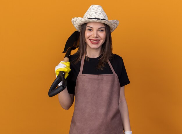 Sorridente giardiniere femmina caucasica che indossa cappello e guanti da giardinaggio che tengono la vanga sulla spalla