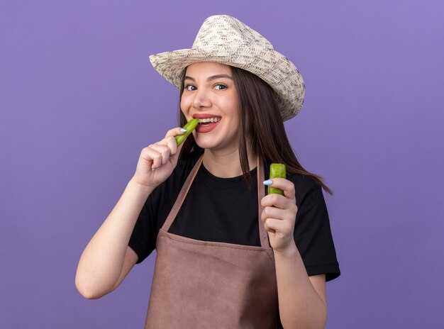 Sorridente giardiniere femmina caucasica che indossa un cappello da giardinaggio che tiene e morde una parte di peperoncino rotto isolato sul muro viola con spazio di copia